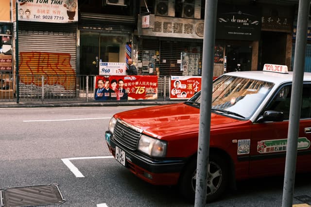 A red taxi parked on a street in an urban area, with graffiti, advertisements, and storefronts in the background