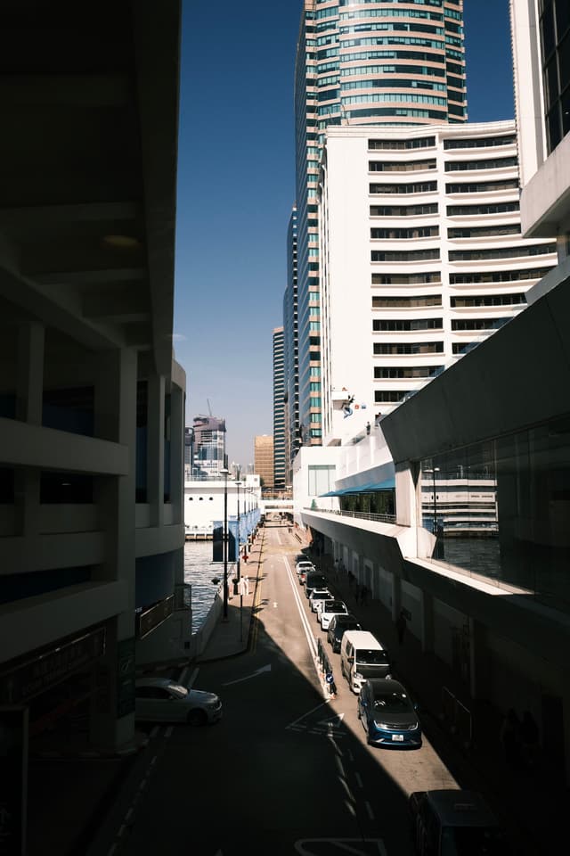 A narrow urban street flanked by modern buildings, with strong sunlight casting sharp shadows