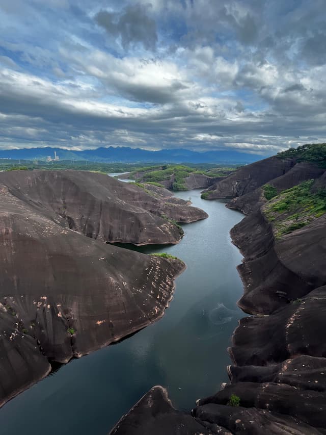 A winding river flows through smooth, dark rock formations under a dramatic cloudy sky, with distant green hills and mountains in the background
