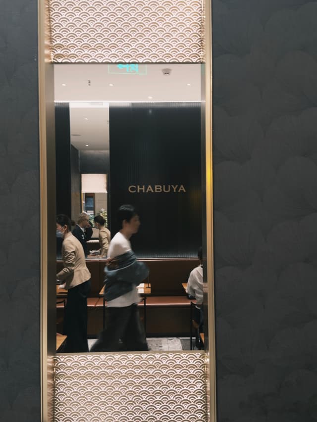 A reflective surface framed with a patterned design shows people walking and dining near a restaurant with the name CHABUYA visible in the background