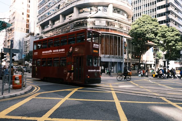 A red double-decker tram moves through a busy urban intersection surrounded by modern buildings and pedestrians