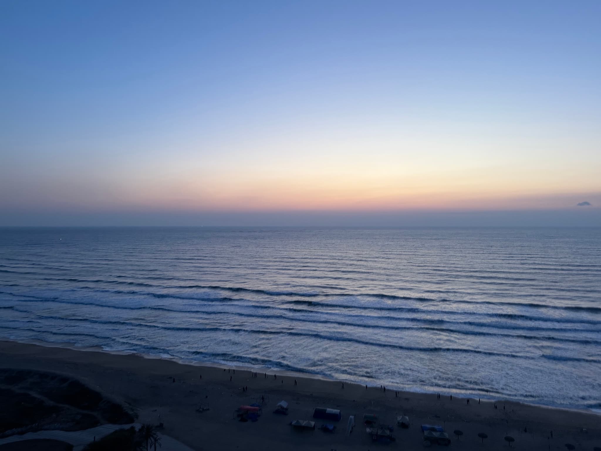 A serene beach at sunset with gentle waves, a clear gradient sky, and parked vehicles along the shore