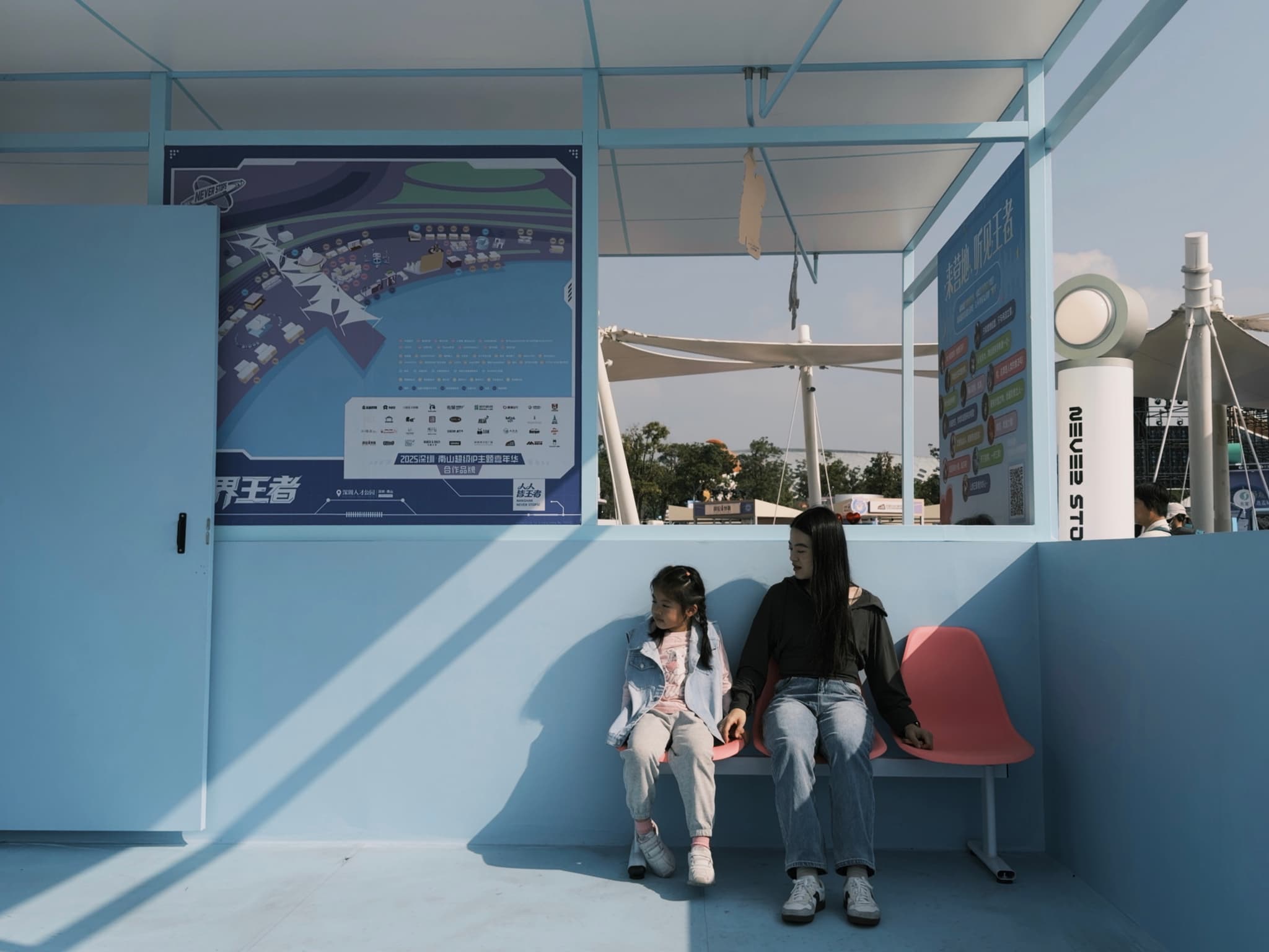Two people sitting on a bench in a light blue structure with maps and signs on the walls, under a partially shaded area