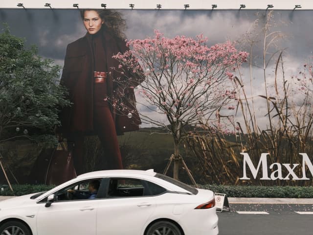 A large fashion billboard featuring a model in a dark outfit with trees and flowers in the background, positioned above a white car on the street