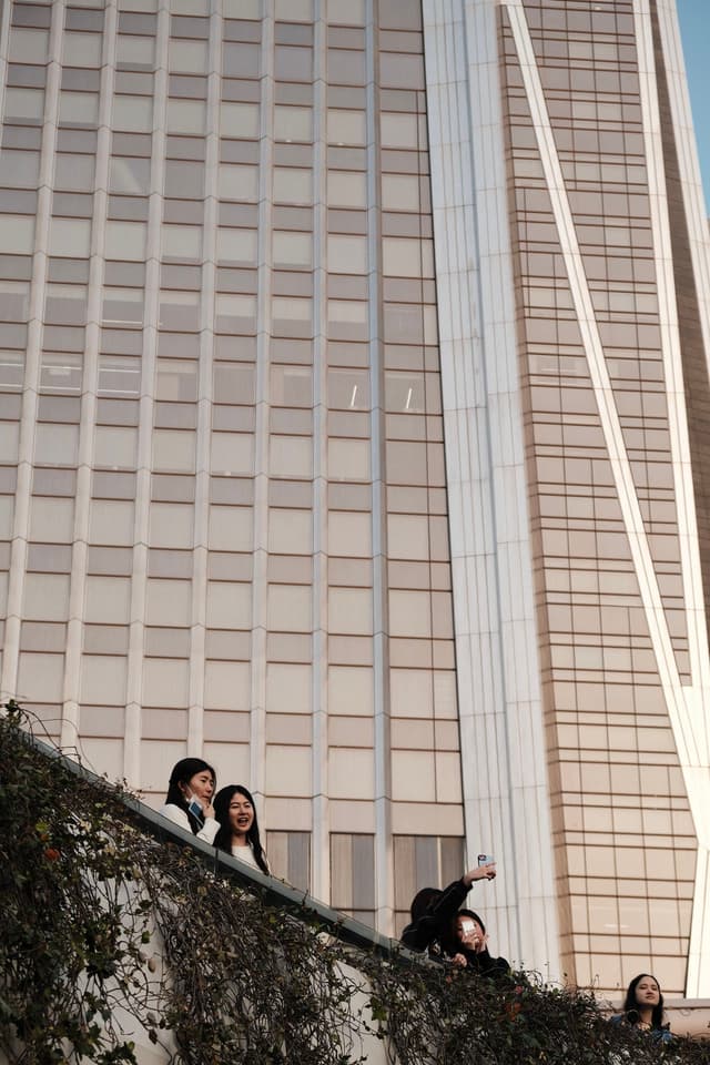 People peeking out from behind a steep rock face with a modern building in the background