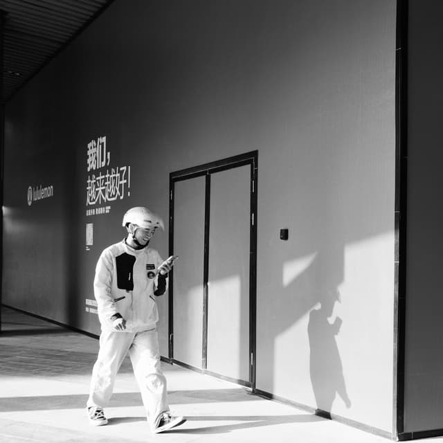 A person wearing a helmet and mask walks while looking at a phone, casting a shadow on a minimalist wall with text and doors