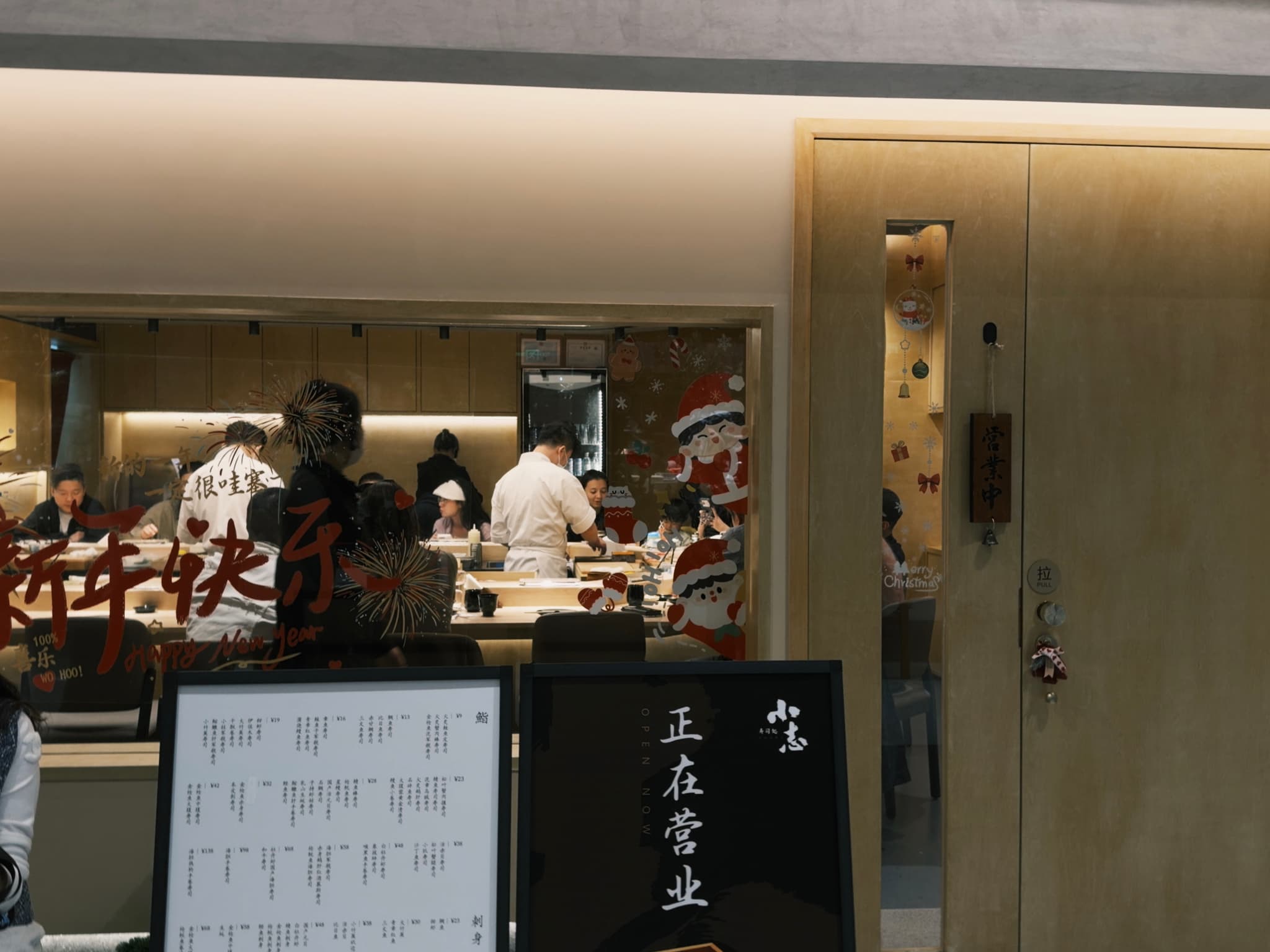 A restaurant interior with chefs working in an open kitchen, a menu board displayed outside, and a wooden door on the right