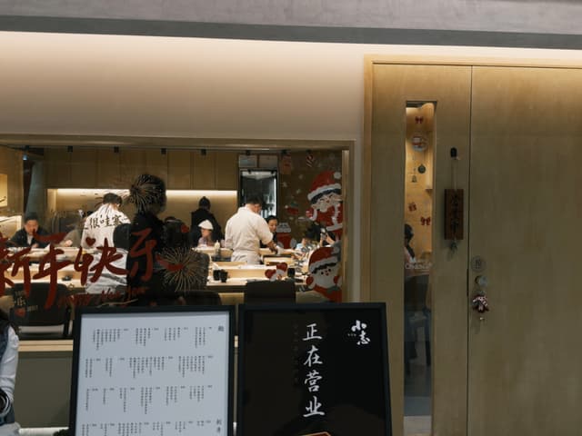 A restaurant interior with chefs working in an open kitchen, a menu board displayed outside, and a wooden door on the right