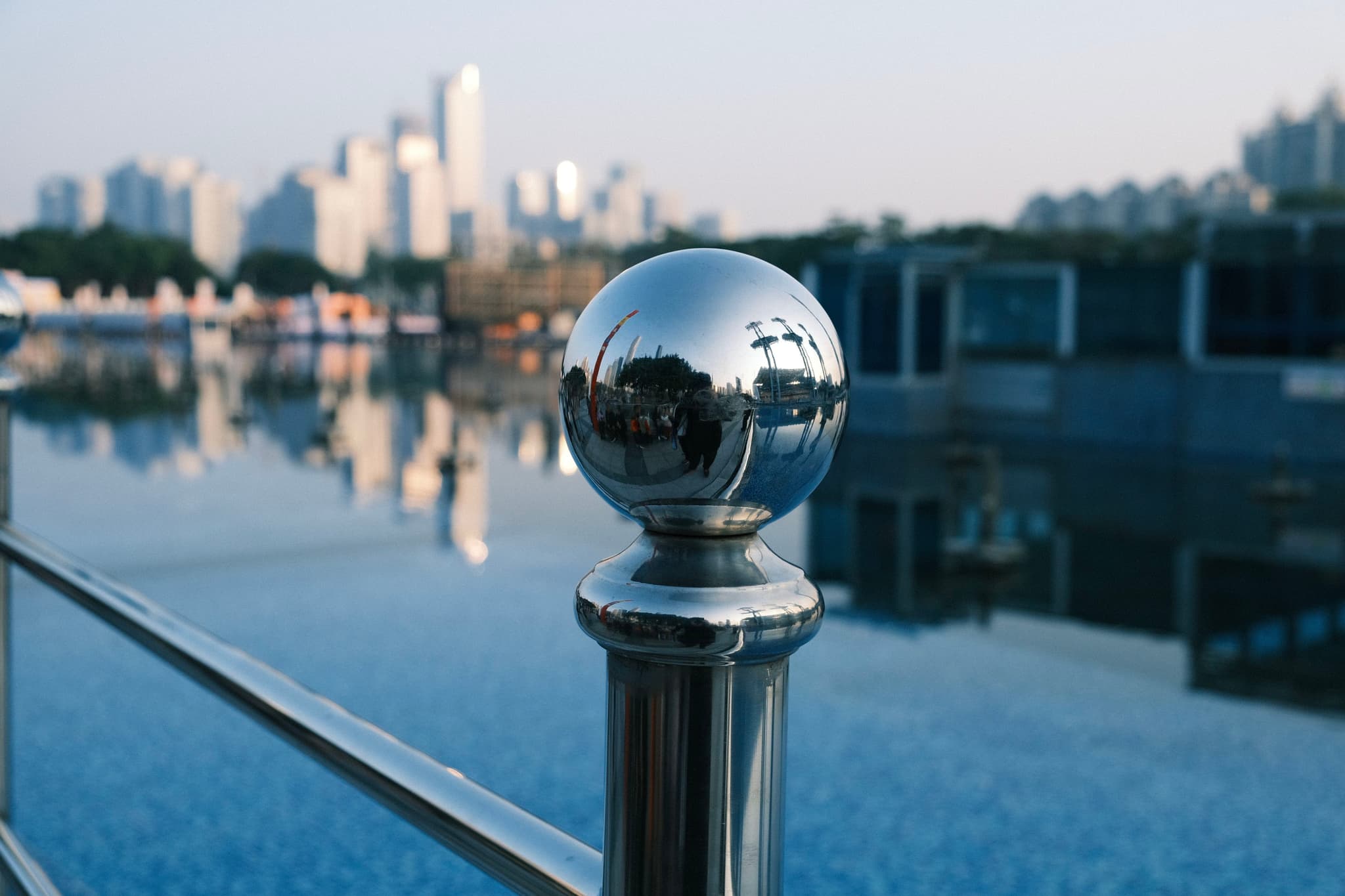 A reflective metal sphere atop a railing with a blurred cityscape and water in the background
